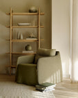 The Ethnicraft Weave Lounge Chair in green linen-blend fabric sits by a light wood shelf with ceramic vases, bowls, and books in a softly lit, minimalist space with neutral tones. Some books are stacked on the floor nearby.
