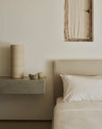 Minimalist bedroom featuring the Ethnicraft Revive Bed - Sand, outfitted with 100% linen removable covers, complemented by crisp white bedding, a concrete shelf displaying neutral-toned pottery, and a small window dressed with a beige curtain set against a light-colored wall.