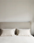 A minimalist bedroom featuring the Ethnicraft Revive Bed - Sand with 100% linen covers, a beige upholstered headboard, white pillows and bedspread, dark round bedside tables, and modern white pendant lights against a light wall.