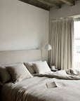 A minimalist bedroom with a neatly made Ethnicraft Revive Bed - Sand in 100% linen, several pillows, an open book, light curtains at the window, and a pendant lamp above a small bedside table.