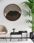 Modern living room with a round abstract wall mirror, tall potted palm, beige armchair, and two black nesting tables with lamp and tray on the Ethnicraft Nomad Kilim Rug - Sand, handwoven from 100% sheep wool.