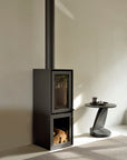 A modern black wood-burning stove with firewood below stands beside the Ethnicraft Oblic Side Table - Black, which holds a cup and saucer. The minimalistic scene features neutral walls and a gray rug.