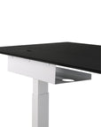 A close-up of a modern sit-stand desk with a black tabletop, white adjustable leg, and an Ethnicraft Metal Cable Tray for organized under-desk cables, set against a white background.