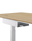 A close-up of a modern desk with a light wood tabletop, white metal legs, and an Ethnicraft Metal Cable Tray for under-desk cable organization. A round grommet hole on the tabletop adds functional European-inspired design.