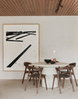 The Ethnicraft Elements Dining Table, paired with four wooden chairs, sits under a striking abstract black-and-white artwork. Minimalist decor, black bowls, and a candle complement the wood-paneled ceiling and neutral microcement walls.