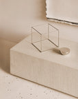 The Elements Floor Shelf by Ethnicraft is showcased with a thin metal wire cube and metallic half-sphere atop a microcement-textured beige block, set against a neutral backdrop with a partially visible canvas.