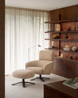 An Ethnicraft Merge lounge chair and matching ottoman on a dark metal swivel base, positioned in a contemporary interior with wood shelving.