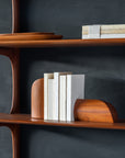 The Ethnicraft PI Wall Shelf - Mahogany (3 shelves) features a few upright white books between two curved solid mahogany bookends, additional closed books, and wooden trays. The shelf is set against a dark textured wall.