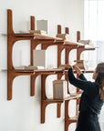 A person arranges neutral-covered PI collection books on the Ethnicraft PI Wall Shelf - Mahogany (2 shelves), styled in a modern mid-century design, as natural light streams in from a window on the right.
