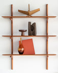 The Ethnicraft PI Wall Shelf - Mahogany (3 shelves) displays modern decor, including abstract sculptures, a brown wooden bowl, a brown glass vase, and a red book positioned at an angle.