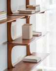 The PI Wall Shelf - Mahogany (5 shelves) by Ethnicraft showcases books upright and stacked against a white wall near a window with sheer curtains, illuminated by natural light.