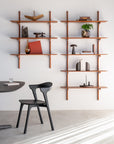 A minimalist interior showcases the Ethnicraft PI Wall Shelf - Mahogany (5 shelves) displaying books, vases, and decor. A modern black chair sits by a round table on a light floor, bathed in sunlight from nearby blinds.