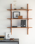 The Ethnicraft PI Wall Shelf - Mahogany (set of 3) displays decor, books, and photos above a modern desk with framed art and a mahogany tray, all arranged against a crisp white wall.