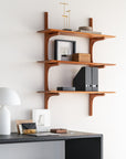 The Ethnicraft PI Wall Shelf - Mahogany (set of 3 shelves) displays decor, books, and boxes above a dark desk with framed photos, a modern white lamp, and small accents against a clean white wall.