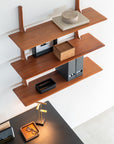 The Ethnicraft PI Wall Shelf - Mahogany (3 shelves) displays decorative boxes above a black desk with a lamp, tray, and glasses, creating a minimalist, modern workspace.