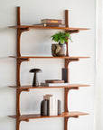 The Ethnicraft PI Wall Shelf - Mahogany (5 shelves) displays books, a potted plant, a small black lamp, and minimalist décor against a white wall illuminated by natural light.