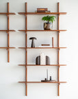 The Ethnicraft PI Wall Shelf - Mahogany (5 shelves) creates an organized, modern display on a white wall, holding a potted plant, books, a black mushroom-shaped lamp, abstract bookends, and decorative objects.