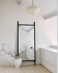 A minimalist room with white walls and light wood floors features the Ethnicraft Pi Floor Mirror - Wide with a black frame, a large white vase with bare branches, modern white cabinets, and a round paper pendant light.