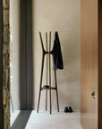 The Pi Coat Stand - Mahogany by Ethnicraft stands in a minimalist room, holding a dark jacket, with black shoes and a small item at its base as sunlight pours through a large window.