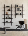 A minimalist living space features Ethnicraft’s Pi Wall Shelf in dark mahogany (5 shelves) displaying books and vases. Below are a modern black chair, floor lamp with a natural shade, and a low table on a textured beige rug.