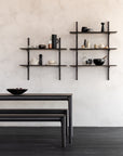 A minimalist kitchen with a black wooden table and the Bok Bench - Brown Oak by Ethnicraft, plus dark shelves displaying bowls, jars, cups, and bottles against a light textured wall.