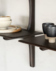 Two dark mahogany Pi Wall Shelves by Ethnicraft display stacked beige bowls and plates on the left, and two black-and-beige PI collection ceramic cups on the right, all set against a light-colored wall.