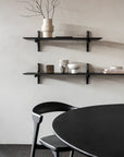 A minimalist dining area features a black round table, matching chair, and the Ethnicraft Pi Wall Shelf in Dark Mahogany displaying neutral vases, bowls, books, and decor against a light-toned wall.
