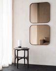 A minimalist room featuring two Ethnicraft Camber wall mirrors in bronze with wooden square frames on a beige wall, a small round dark wood table with a white cylindrical lamp and magazines, and sheer white curtains by the window.