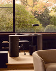A modern home office features the Ethnicraft Roller Max Desk, an office chair, a black desk lamp, and a large window revealing lush greenery and autumn leaves. A beige armchair sits in the foreground.