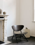 An Ethnicraft Eye Lounge Chair in mahogany sits in the corner of a minimalist room with white walls, near a marble fireplace. Next to it, a small round side table holds a teacup, saucer, and stacked books.