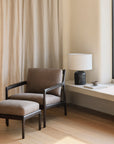 The Jack Lounge Chair - Nubuck Leather by Ethnicraft sits on wooden flooring next to a white table, showcasing Scandinavian modernism against a beige curtain and cream wall.