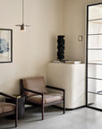 Minimalist room with two Ethnicraft Jack Lounge Chair - Nubuck Leather, a small side table, framed wall art, sculpture on a white pedestal, and glass door—decor reflecting Scandinavian modernism in neutral tones.