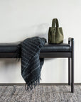 A green handbag and a dark, fringed striped blanket from Ethnicraft rest on the Jack Bench - Mahogany with Black Leather against a light gray wall, with part of a textured rug visible on the floor.