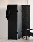 The Ethnicraft Chopped Room Divider in black stained mahogany stands in a minimal room, one panel draped with a black garment, while stacked books and a black chair rest nearby on a light-colored floor.