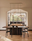 Modern dining room with an Ethnicraft Sequence Dining Table in dark mahogany, two chairs, a light accent chair, decorative bowls, sculptural glass pendants, large window with treetop views, parquet flooring, and warm neutral walls.