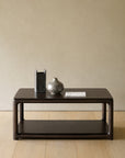 A dark wood Ethnicraft Eda Coffee Table | Square with a lower shelf, topped with a rectangular vase, a round vase, and a book.
