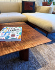 A colorful book rests on the SOBU Bolinas Coffee Table in Claro Walnut, placed before a beige sectional with a dark cushion. The modern room includes a woven rug and a potted plant in the corner.