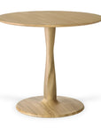 The Torsion Dining Table by Ethnicraft features a round oak top with a natural finish, refined woodworking, and a single central pedestal leg that curves gently into a wide circular base.