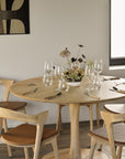 The Ethnicraft Torsion Dining Table - Oak, set for four with glassware, cutlery, and a floral centerpiece, pairs with wooden chairs featuring brown cushions in a bright, modern minimalist room.