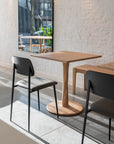 A modern cafe interior featuring Ethnicraft’s Torsion Square Dining Table in oak, two black chairs, a bench, white brick walls, and a large window streaming natural light.