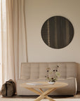 A minimalist living room features a beige loveseat, the Ethnicraft Mikado Coffee Table Round in solid oak, round dark wall art, full-length curtains, and a brown bag on the floor. Natural light fills the space.