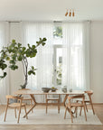 A modern dining room features a wooden table with four chairs on the Nomad Kilim Rug - Sand by Ethnicraft, crafted from 100% sheep wool. A tall potted plant stands in the corner, and sheer white curtains cover large windows.