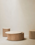 Three Ethnicraft Roller Max Coffee Tables in solid oak, each at a different height with vertical grooves, are displayed on a light patterned floor against a plain beige wall for a minimalist modern look.