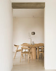 Minimalist dining area featuring the Ethnicraft Roller Max Round Dining Table in oak, six wooden chairs, a pendant light overhead, and a vase with dried flowers. Light walls and floors create a calm, airy atmosphere.