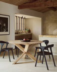 A modern dining area featuring the Ethnicraft Circle Dining Table in European oak, surrounded by four contract-grade black chairs and accented with a fruit bowl centerpiece. Natural wood and stone elements complement the space, while an Ethnicraft black-and-white photo adorns the wall near the open kitchen.