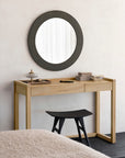 An Ethnicraft Frame Desk - Oak sits against a light wall, paired with a black stool and topped with decorative boxes and a small dish. Above, a large round mirror with a textured dark frame is mounted.