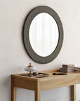 A round mirror with a textured, cracked frame hangs above an Ethnicraft Frame Desk - Oak, which displays decorative boxes, a lidded glass dish, and a makeup brush, all set against a light-colored wall.