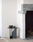 A minimalist interior features a white wall, stone fireplace, and the Ethnicraft Geometric Side Table in black oak. The light wood floor complements a black bowl and dish, with firewood stored in the fireplace.