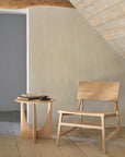A minimalist room features Ethnicrafts N2 Lounge Chair in oak and a matching side table, holding books and a coffee cup, set against textured beige walls with wooden floors under a sloped ceiling.