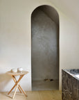 Minimalist interior with a light wooden floor, Ethnicrafts Mikado Side Table in oak displaying two white accents, a marble bathtub, and an arched doorway opening to a simple gray alcove.